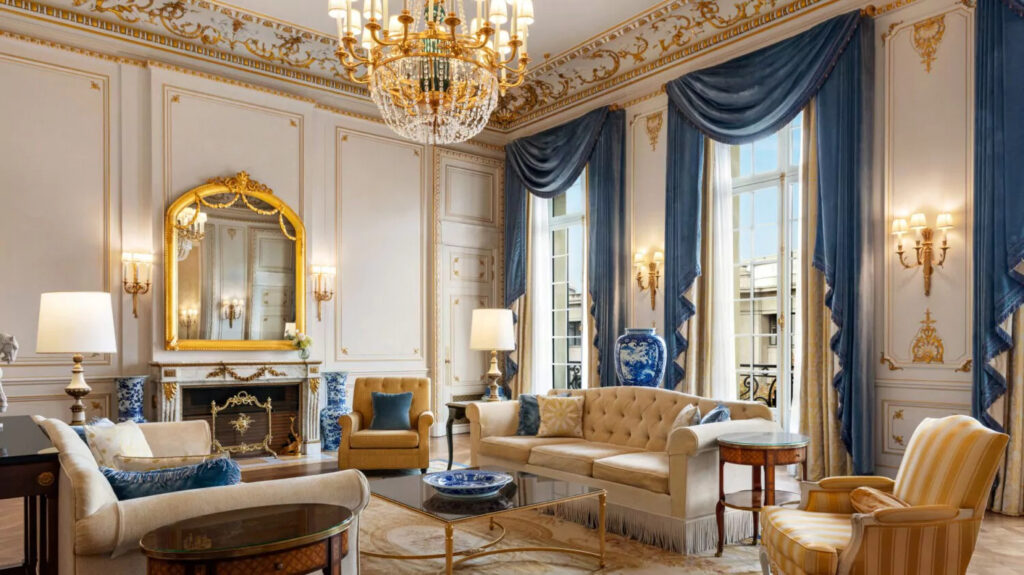 Un salon luxueux de style classique avec des moulures dorées, des rideaux en velours bleu, un lustre imposant, des meubles élégants et une cheminée ornée d'un miroir doré." 