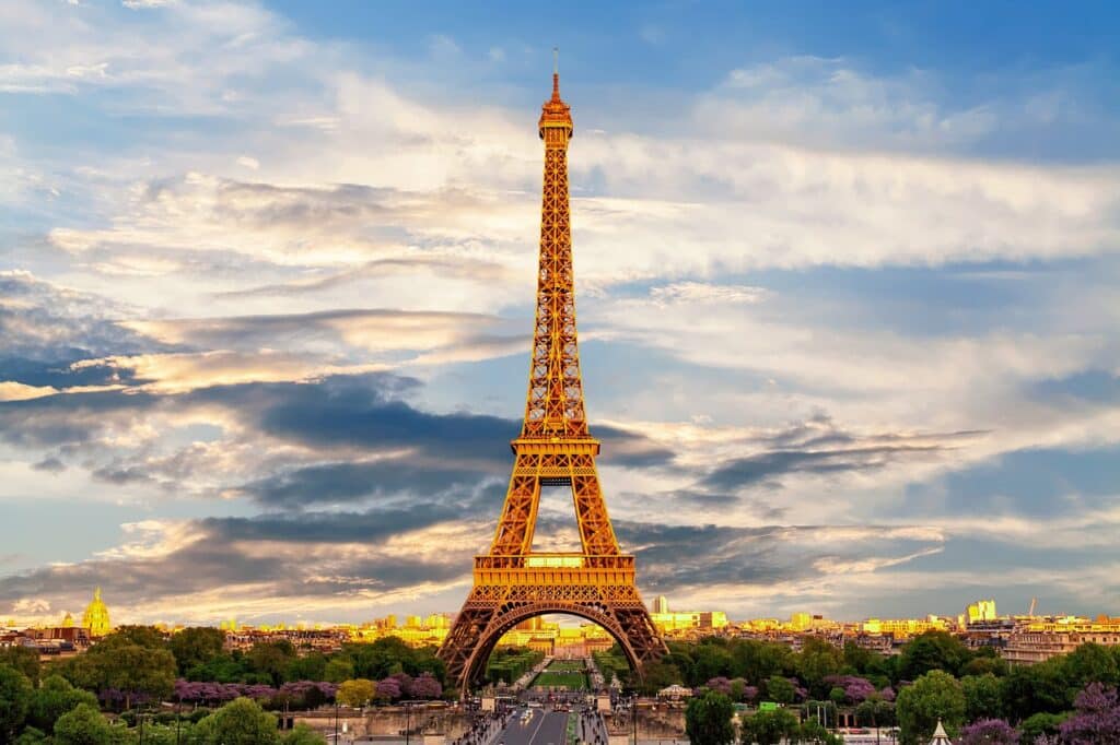 La Tour Eiffel illuminée par les rayons du soleil couchant, entourée de verdure et surplombant un ciel partiellement nuageux aux teintes dorées et bleutées." 