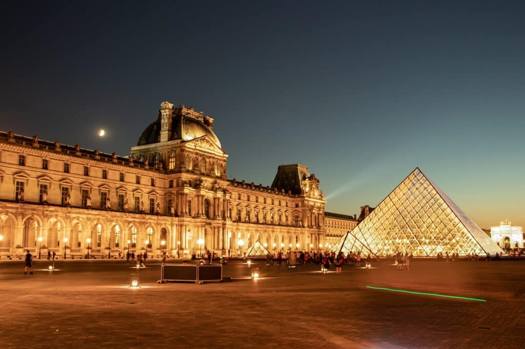 Vue nocturne du musée du Louvre à Paris, illuminé avec sa célèbre pyramide en verre scintillante, sous un ciel dégagé avec un croissant de lune visible à l'horizon