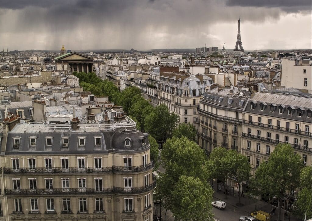 Vue panoramique sur Paris avec la Madeleine au premier plan, des immeubles haussmanniens bordés d'arbres, et la Tour Eiffel se dressant sous un ciel nuageux